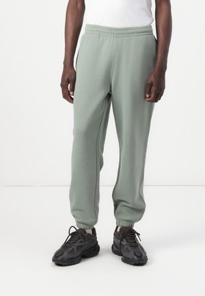 Homme portant un pantalon de jogging vert sauge et des chaussures de sport noires, debout sur un fond blanc.