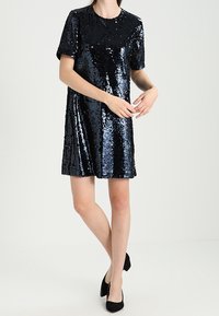 Kvinna klädd i en kortärmad, knälång svart paljettklänning med svarta klackar, stående med benen korslagda och händerna försiktigt rörande vid varandra.