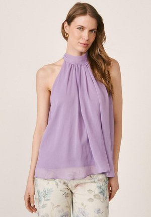 Mujer con cabello largo y castaño que lleva pendientes de aro dorados, una blusa lila sin mangas con cuello halter y pantalones claros con estampado floral, de pie frente a un fondo sencillo.