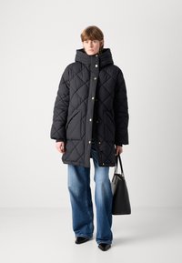 Zwarte gewatteerde pufferjas met een capuchon, voorzien van gouden drukknopen en twee frontzakken, gecombineerd met een wijde jeans in het blauw en een zwarte handtas.