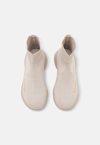 Beige klouzací tenisky s ribbovaným kotníkem, flexibilním pleteným materiálem a texturovaným povrchem. Obsahuje zlatý akcent loga.