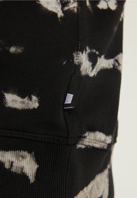 Gros plan sur un tissu noir avec un motif tie-dye blanc et une couture visible, avec une petite étiquette rectangulaire noire et blanche.