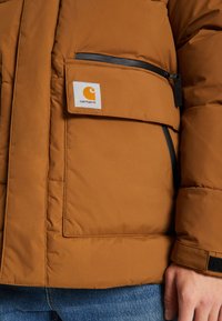 Brun pufferjakke med en blød tekstur, har en fremtrædende frontlomme og sort lynlås, prydet med et Carhartt logo patch.