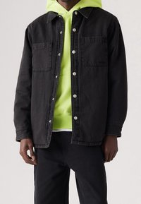 Černá džínová bunda se zapínáním na patentky, dvě náprsní kapsy, nošená přes neonově zelenou mikinu s kapucí, v kombinaci s černými kalhotami, zobrazená zepředu.