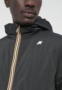 Chaqueta ligera negra con capucha, que presenta una cremallera multicolor y un logo blanco en el pecho. Tejido suave y repelente al agua.