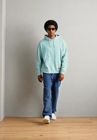 Felpa con cappuccio azzurro chiaro e lacci bianchi, abbinata a jeans blu e sneakers bianche. Il modello indossa occhiali da sole scuri.