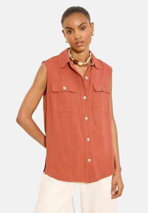 Kobieta w bezrękawnym, rdzawego koloru koszuli zapinanej na guziki z dwoma kieszeniami na piersi, w połączeniu z białymi spodniami i masywnym naszyjnikiem z koralików.