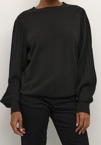 Person in einem schwarzen Pullover mit gefalteten Ballonärmeln und schwarzen Hosen, stehend vor einem schlichten hellen Hintergrund.