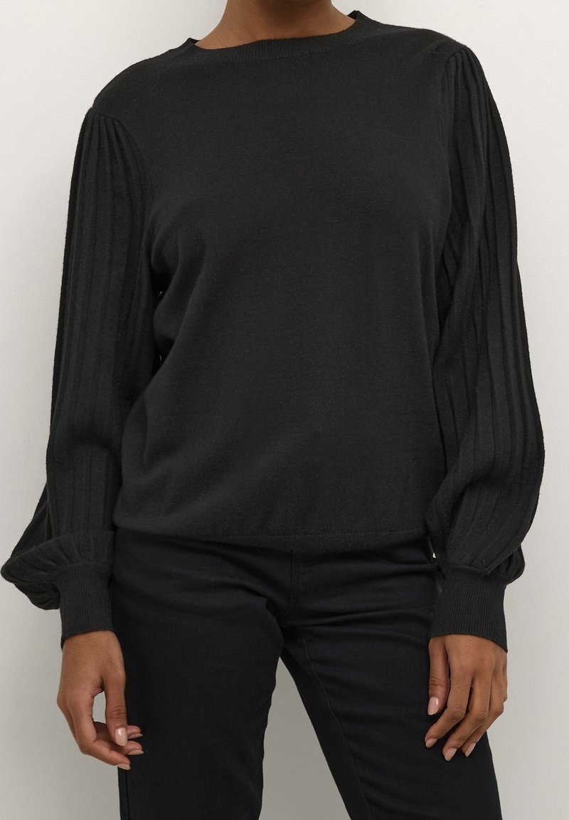 Person in einem schwarzen Pullover mit gefalteten Ballonärmeln und schwarzen Hosen, stehend vor einem schlichten hellen Hintergrund.