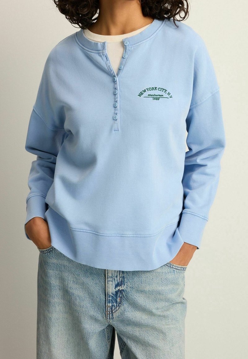 Personne portant un sweat-shirt bleu clair boutonné avec le texte "New York City N.Y. Manhattan 1985" et un jean bleu clair, mains dans les poches.