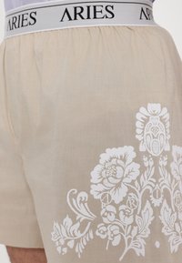 Boxer in beige con un motivo floreale bianco e cinturino con il marchio "ARIES". Il tessuto è leggero e ha una consistenza liscia.
