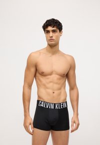 Crne Calvin Klein bokserice s širom bijelom elastičnom trakom, izrađene od glatkog materijala, s uskim oblikom i rezom u stilu gaćica.