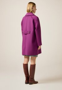 Lila Wolljacke mit großem Kragen, Rückentaschendetail und zwei Knöpfen. Braune Lederstiefel bis zur Wade und sichtbarer karierter Rock.