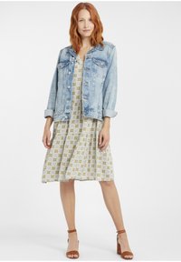 Jachetă din denim albastru deschis cu accesorii argintii, purtată peste o rochie cu model galben și alb, având un design în straturi și sandale cu toc maro.