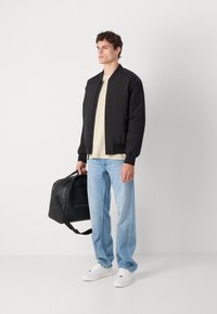 Black bomber jacket, beige t-shirt, light blue jeans, and white sneakers. Holds a black bag with short handles and a detachable shoulder strap.