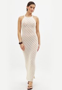 Maxi dress in tessuto a maglia crema, con un motivo a onde, design senza maniche e fessure laterali. Abbinata a un bracciale marrone e sandali.