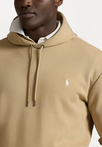 Beige hoodie van een gestructureerde stof, met een wit logo op de borst, trekkoorden en een gelaagd kraagontwerp.