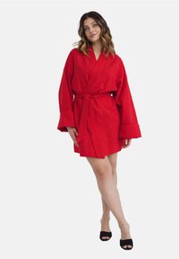 Czerwony lniany szlafrok z długimi rękawami i dopasowanym paskiem, o luźnym kroju i teksturowanej powierzchni. Widoczne czarne buty z odkrytymi palcami.