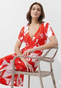 Rotes florales Wickelkleid mit kurzen Ärmeln, verfügt über weiße Akzente und einen tiefen V-Ausschnitt. Sitzt auf einem hellen Stuhl. Glatte Stoffstruktur.