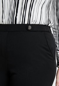Pantalon noir à la texture lisse, avec une fermeture à bouton et des poches latérales. Associé à une chemise blanche à rayures verticales noires.