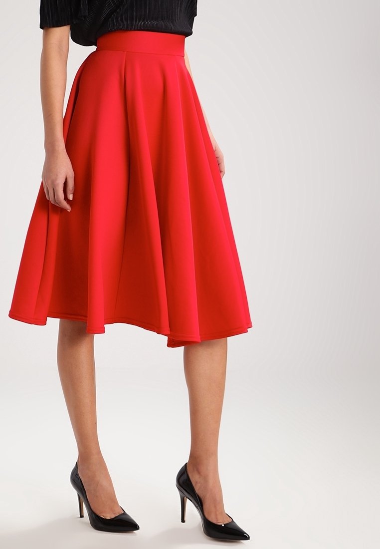 Femme portant une jupe évasée rouge vif jusqu'aux genoux, un haut noir et des chaussures à talons hauts noires, debout devant un fond uni.