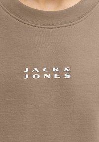Lähedalt vaade beežile kangast särgile, millel on valge "JACK & JONES" logo trükitud kaeluse alla keskele.