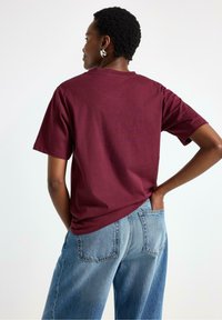 Mørkerød bomulds T-shirt med korte ærmer, afslappet pasform; båret med højtaljede lyseblå jeans, der viser rygdesignet og tekstur.