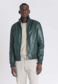 Groene leren bomberjack met een hoge kraag, geribde manchetten en zoom, rits aan de voorkant en zijzakken. Model draagt een lichtgekleurde broek.