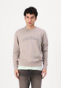 Felpa marrone chiaro con il testo "BELSTAFF" in un design curvo. Presenta un colletto rotondo e polsini a costine, indossata sopra una maglietta verde menta.