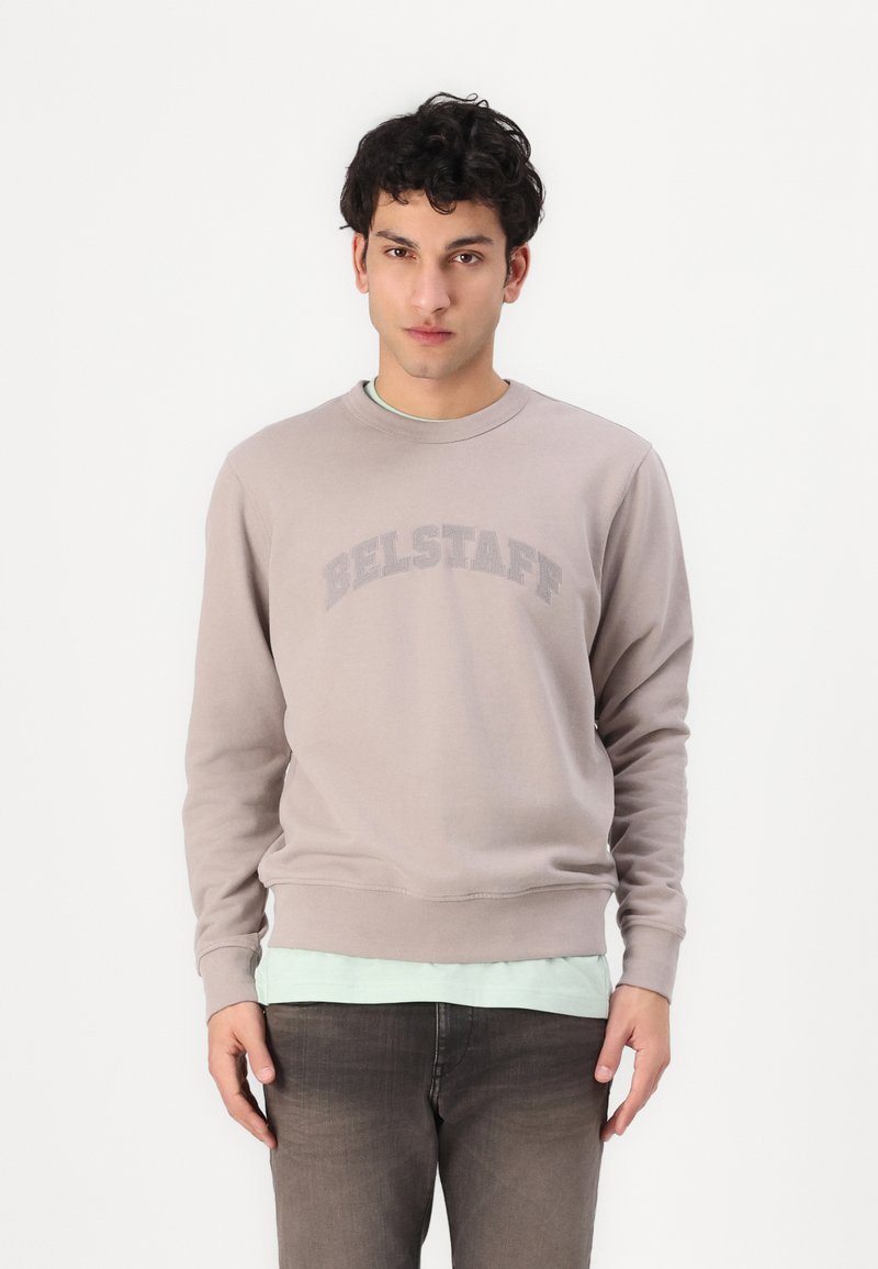 Felpa marrone chiaro con il testo "BELSTAFF" in un design curvo. Presenta un colletto rotondo e polsini a costine, indossata sopra una maglietta verde menta.