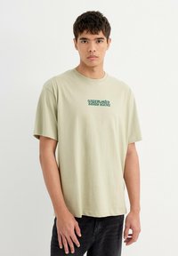 Camiseta de algodón de manga corta en verde claro. Presenta un texto gráfico de punto "Ciencia de Botánica Submarina" en la parte delantera. Corte simple y relajado.
