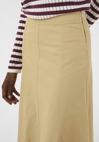 Jupe longue beige avec fermeture éclair sur le côté, fabriquée en tissu durable. Présente des lignes épurées et une texture subtile. La main du mannequin repose sur la jupe.