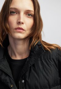 Junge Frau mit hellbraunem Haar trägt eine schwarze gesteppten Jacke mit vertikalen Nietenstreifen vor einem schlichten hellen Hintergrund.