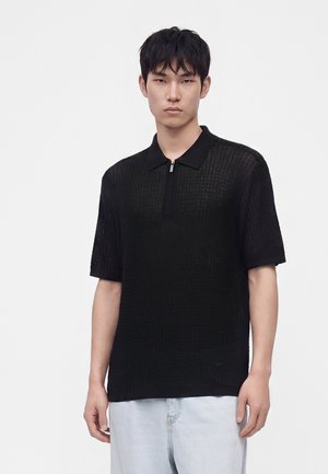 Jeune homme portant un pull en maille noir à manches courtes avec un col zippé et un jean bleu clair sur fond blanc uni.