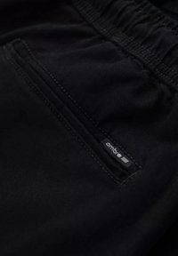 Primer plano de un bolsillo de shorts de tela negra con costuras visibles y pequeña etiqueta de marca "ombre" cerca de la cintura elástica.