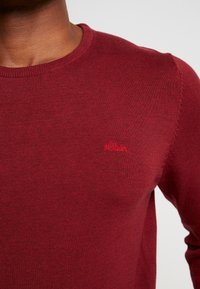 Rød strikket sweater med rund halsudskæring og ribbede kanter. Har et lille, broderet s.Oliver-logo på brystet. Glat tekstur.