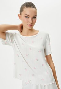 Vit kortärmad topp med ribbad textur, prydd med rosa blommönster och rundad halsringning. Nederdelen har en lätt scallopad kant.