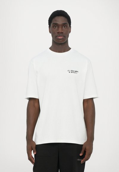 T-shirt en coton blanc avec un col rond et des manches courtes. Présente un imprimé en texte noir sur le côté gauche. Hem droit.