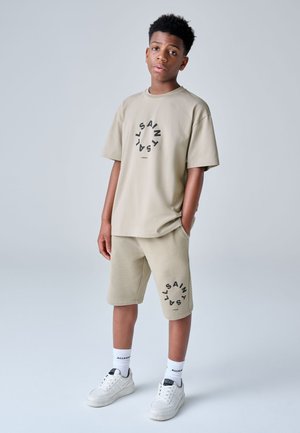 Boy wearing beige matching t-shirt and shorts with circular black text, white socks, and white sneakers, standing against a plain background.
