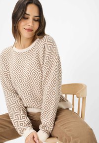 Maglione lavorato a maglia con un motivo geometrico a diamante nei toni del beige e del marrone. Polsini e orlo a coste, texture morbida, vestibilità rilassata. Indossato con pantaloni in velluto a coste.