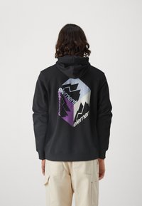 Sweat à capuche noir avec un grand motif graphique au dos présentant un design géométrique et des motifs de montagnes en tons violet, bleu et blanc.