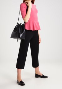 Rosa Peplum-Oberteil mit Dreiviertelärmel, kombiniert mit schwarzen, verkürzten Hosen. Eine schwarze Bucket-Bag hängt über der Schulter, und glänzende schwarze Loafers.