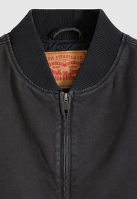 Μαύρο μπουφάν τύπου bomber με ριμπ στο κολάρο, με μπροστινό φερμουάρ και υφασμάτινη ετικέτα που δείχνει το λογότυπο και τις οδηγίες φροντίδας. Λείο υλικό.