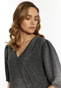 Schwarzes, glänzendes Kleid mit einem V-Ausschnitt, kurzen Puffärmeln und einem strukturierten Stoff. Das Design weist einen subtilen Glitzereffekt auf.