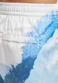 Detail pasu a boků v bílých šortkách s modrými abstraktními vzory a elastickým pasem s kapsou na zadní straně.
