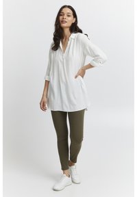 Weißes, langärmliges Tunika mit subtilem Textur und Kragen, kombiniert mit olivgrünen Slim-Fit-Hosen und weißen Sneakers.