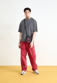 T-shirt grigio a maniche corte, pantaloni rossi larghi, borsa a tracolla nera con portachiavi decorativo, scarpe da ginnastica bianche, in piedi su un pavimento giallo.
