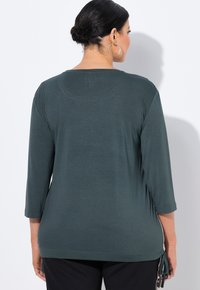 Top de manga larga en verde oscuro con escote redondeado. Textura suave, corte holgado y lazos ajustables en los laterales. Dobladillo ligeramente curvado, diseño simple.