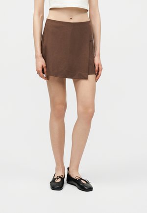 Kobieta w brązowej mini spódnicy z asymetrycznym przodem i czarnych balerinach, stojąca na tle jednolitego, jasnego tła.