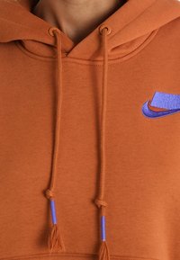 Rostfärgad hoodie i mjukt tyg, med en blå Nike-logotyp och kontrasterande blå tofsar på dragsnörena.